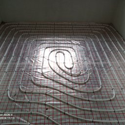 Michał Kulwicki Usługi hydrauliczne - Białe rury ogrzewania podłogowego ułożone spiralnie na folii z czerwoną siatką, przymocowane czarnymi uchwytami, widok z góry.