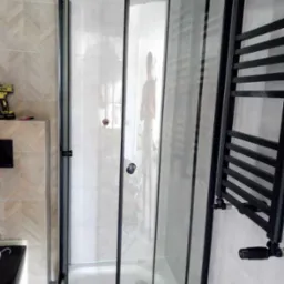 Narożna kabina prysznicowa z czarnymi profilami i grzejnikiem drabinkowym w łazience z beżowymi płytkami, widoczny żółty wkrętak na szafce.