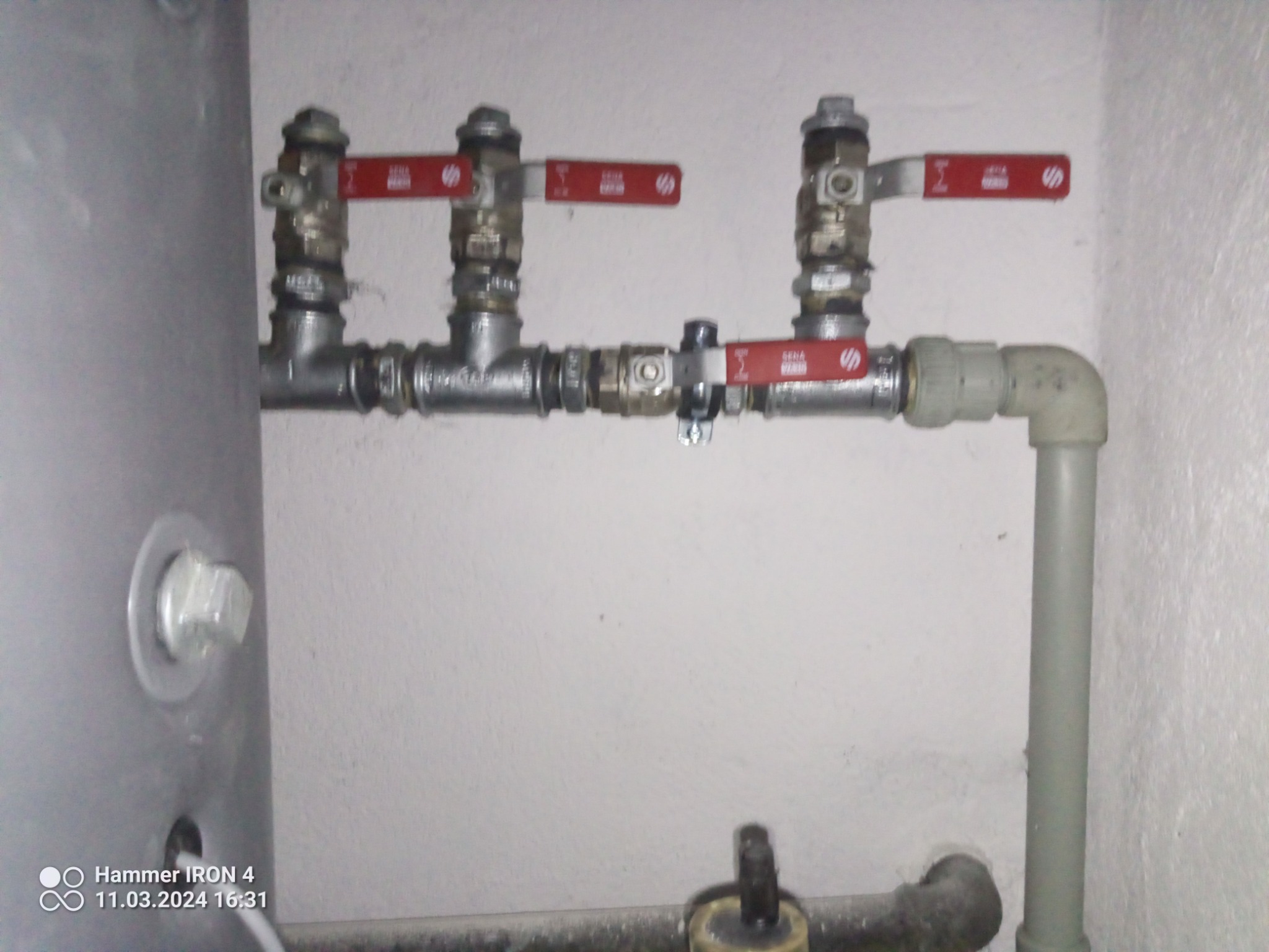 Szare rury instalacji grzewczej z czerwonymi zaworami, widoczne elementy bojlera i ściana z drobnymi zabrudzeniami.
