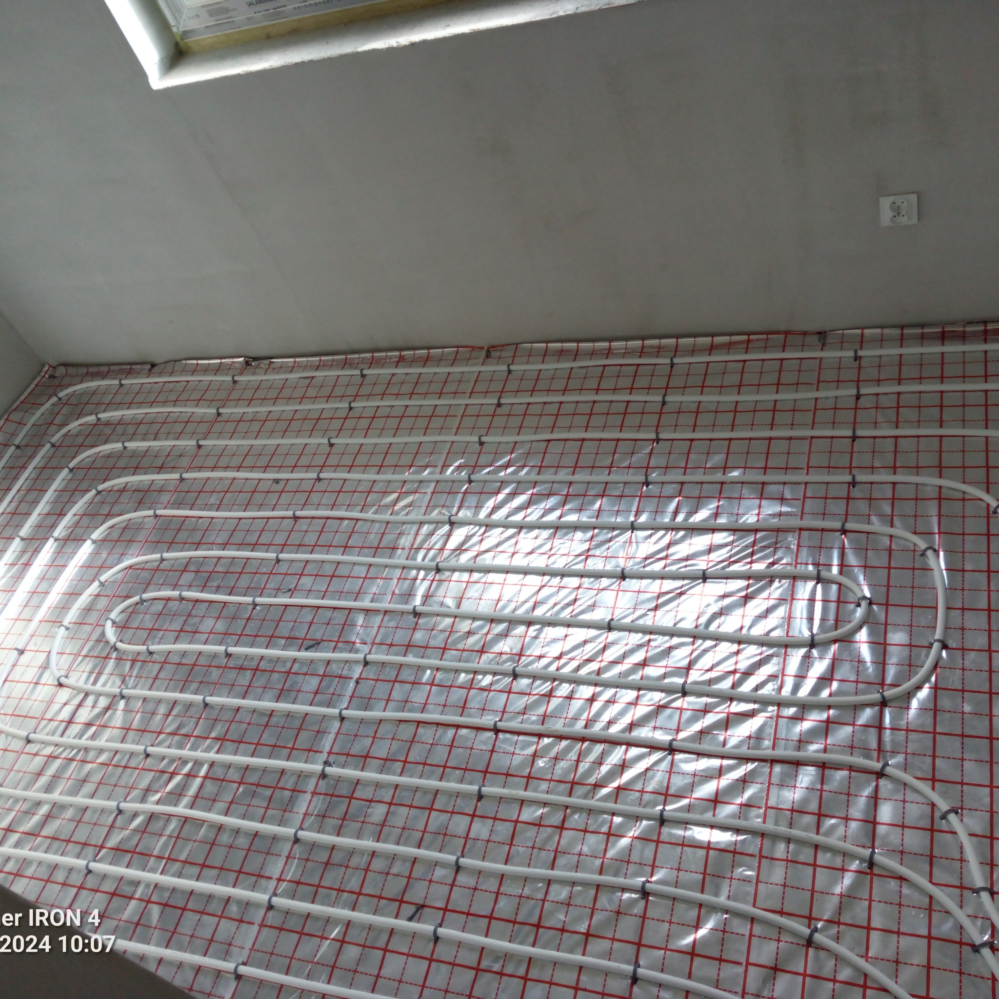 Białe rury ogrzewania podłogowego ułożone na folii z czerwoną siatką na przygotowanym podłożu, widok z góry, surowy stan wykończenia wnętrza.