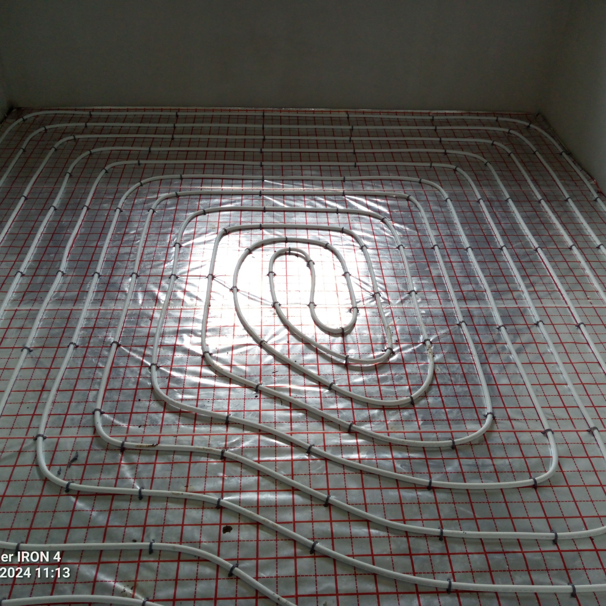 Białe rury ogrzewania podłogowego ułożone spiralnie na folii z czerwoną siatką, przymocowane czarnymi uchwytami, widok z góry.