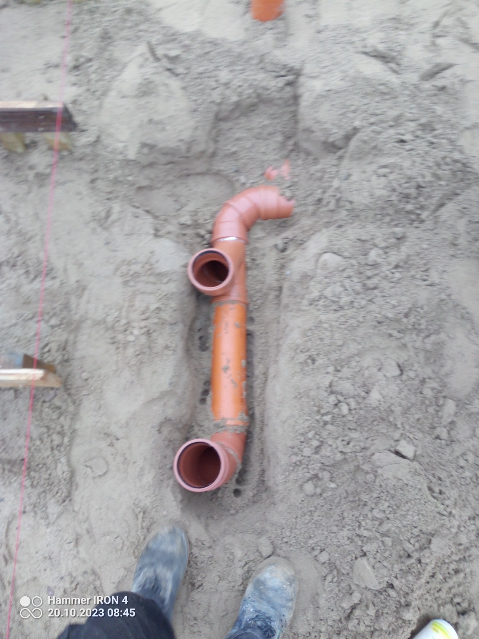 Pomarańczowa rura kanalizacyjna z trójnikiem ułożona w wykopie, widoczne buty pracownika i fragment drewnianej konstrukcji szalunkowej.