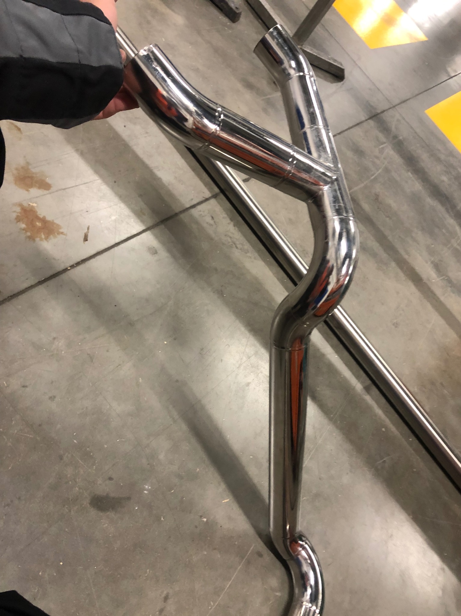 Element konstrukcyjny ze stali nierdzewnej, prawdopodobnie fragment poręczy lub balustrady, z widocznymi spawami i odbiciami, trzymany w dłoni, na tle betonowej podłogi z żółtymi pasami.