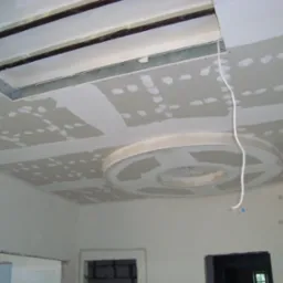 Sufit podwieszany z elementami konstrukcyjnymi i okrągłą dekoracją centralną, w trakcie szpachlowania, widoczne przewody elektryczne, surowy stan wykończenia wnętrza.