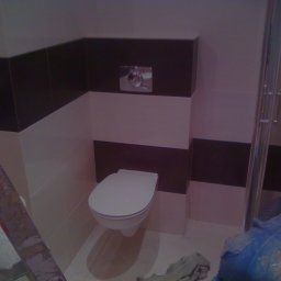 Almark - Biało-brązowa łazienka z wiszącą toaletą, widoczna drabina malarska i fragment sylwetki osoby w niebieskim kombinezonie podczas prac wykończeniowych.