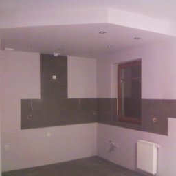 Almark - Wnętrze w Ciechanowie w trakcie prac przygotowawczych do wykończenia kuchni: częściowo położone płytki nad blatem, widoczne wyprowadzenia instalacji elektrycznej i wodnej, grzejnik i okno.