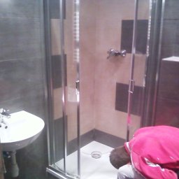 Almark - Montaż kabiny prysznicowej z chromowanymi profilami w łazience z szarymi i beżowymi płytkami, widoczny pracownik w różowej bluzie pochylony nad brodzikiem.