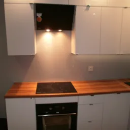 Nowoczesna kuchnia z białymi szafkami, drewnianym blatem i czarną płytą indukcyjną oraz piekarnikiem. Nad płytą znajduje się czarny okap z włączonym oświetleniem punktowym.