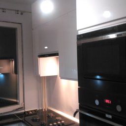 Almark - Nowoczesna kuchnia z białymi, lakierowanymi szafkami, czarnym blatem, płytą grzewczą i piekarnikiem z mikrofalówką w zabudowie, oświetlona wiszącą żarówką i listwą LED pod szafkami.