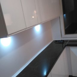 Almark - Nowoczesna kuchnia z białymi, błyszczącymi szafkami górnymi i ciemnym, marmurkowym blatem roboczym, oświetlonym punktowymi lampkami LED pod szafkami, widok na fragment okna.