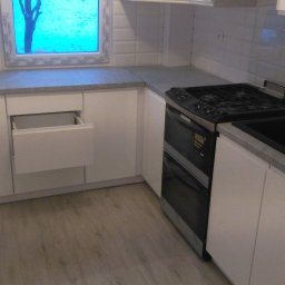 Almark - Nowoczesna zabudowa kuchenna z białymi szafkami, szarym blatem i czarnym zlewem oraz kuchenką. Jedna z szuflad jest otwarta, a w tle widoczne okno z widokiem na drzewo.