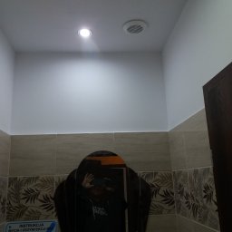 Almark - Wykończona łazienka z beżowymi płytkami z motywem roślinnym, okrągłe lustro odbija osobę robiącą zdjęcie, biały sufit z punktowym oświetleniem i wentylacją, fragment ciemnych drzwi po prawej.