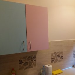 Almark - Narożne szafki kuchenne w pastelowych odcieniach (błękit i róż) z metalowymi uchwytami, zawieszone nad blatem z beżowymi płytkami dekorowanymi motywem liści.