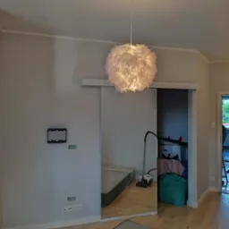 Szare wnętrze pokoju z przesuwnymi drzwiami lustrzanymi, odsłaniającymi materac, odkurzacz i inne przedmioty. Na ścianie widoczna ramka pod telewizor i gniazdka elektryczne, a z sufitu zwisa...