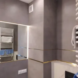 Nowoczesna łazienka wykończona szarymi płytkami z poziomymi, złotymi akcentami; widoczne lustro z podświetleniem LED, grzejnik drabinkowy z ręcznikami oraz fragment ściany z włącznikiem światła...