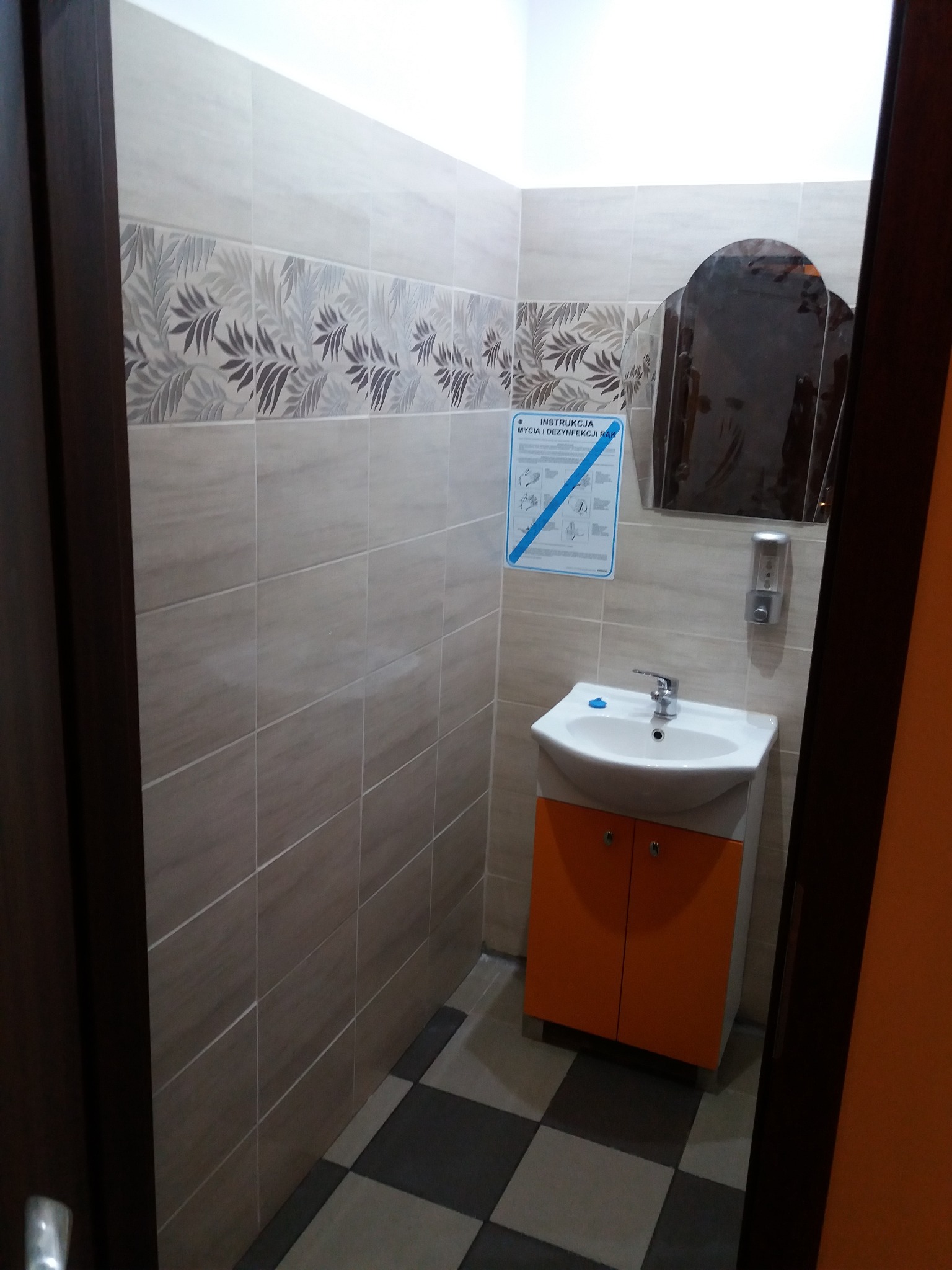 Wykończona łazienka z beżowymi płytkami z motywem roślinnym, białą umywalką z pomarańczową szafką, instrukcją mycia rąk i lustrem w drewnianej ramie.