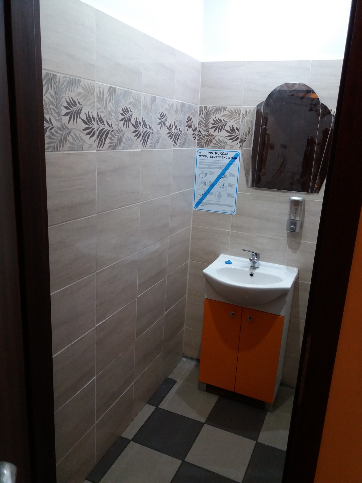 Umywalka z pomarańczową szafką w łazience wyłożonej beżowymi płytkami z dekoracyjnym pasem z motywem liści, na ścianie instrukcja mycia rąk i dozownik mydła