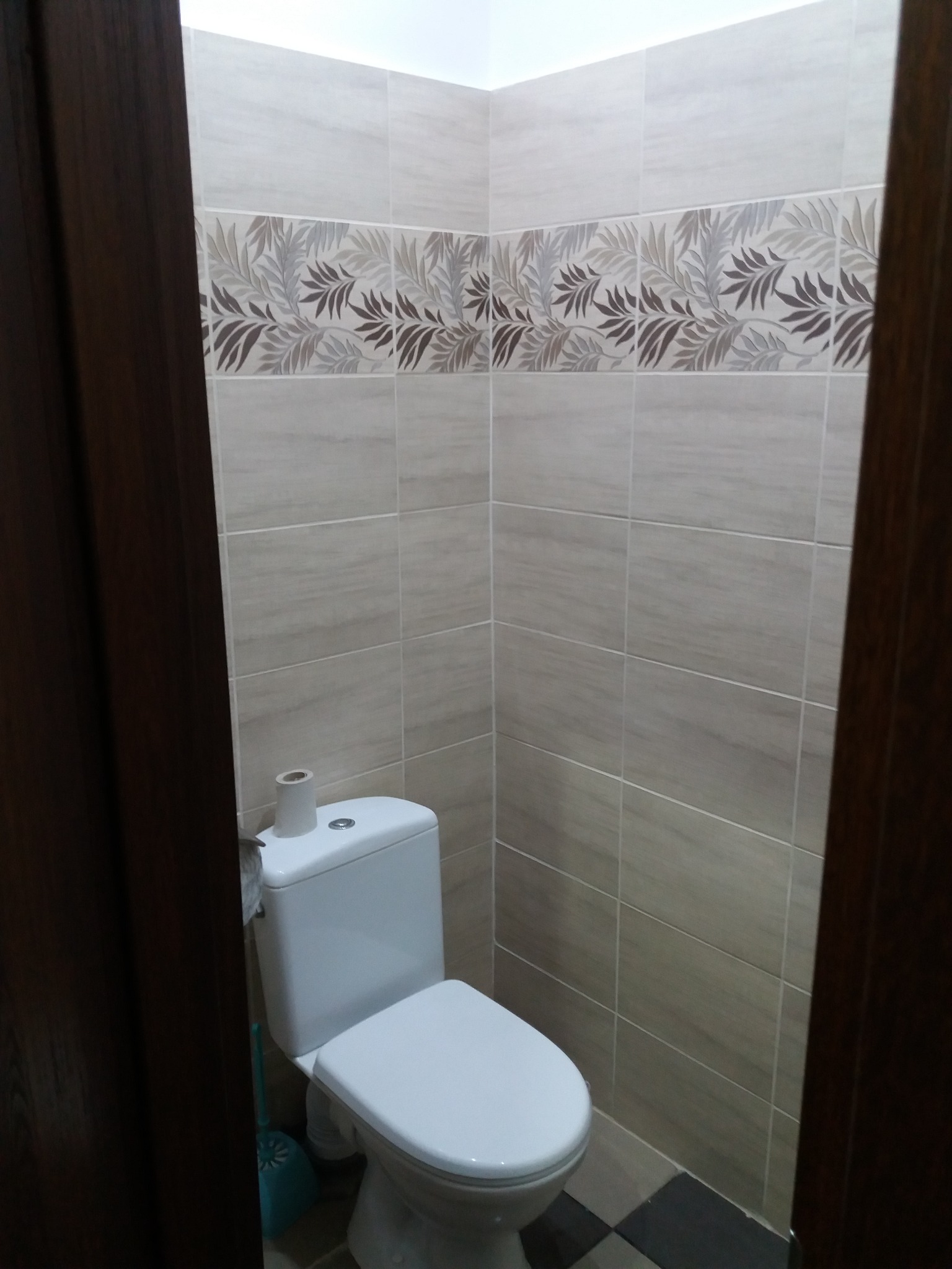 Wykończona toaleta z beżowymi płytkami, dekoracyjnym pasem z motywem liści i ciemnobrązowymi drzwiami po bokach kadru.