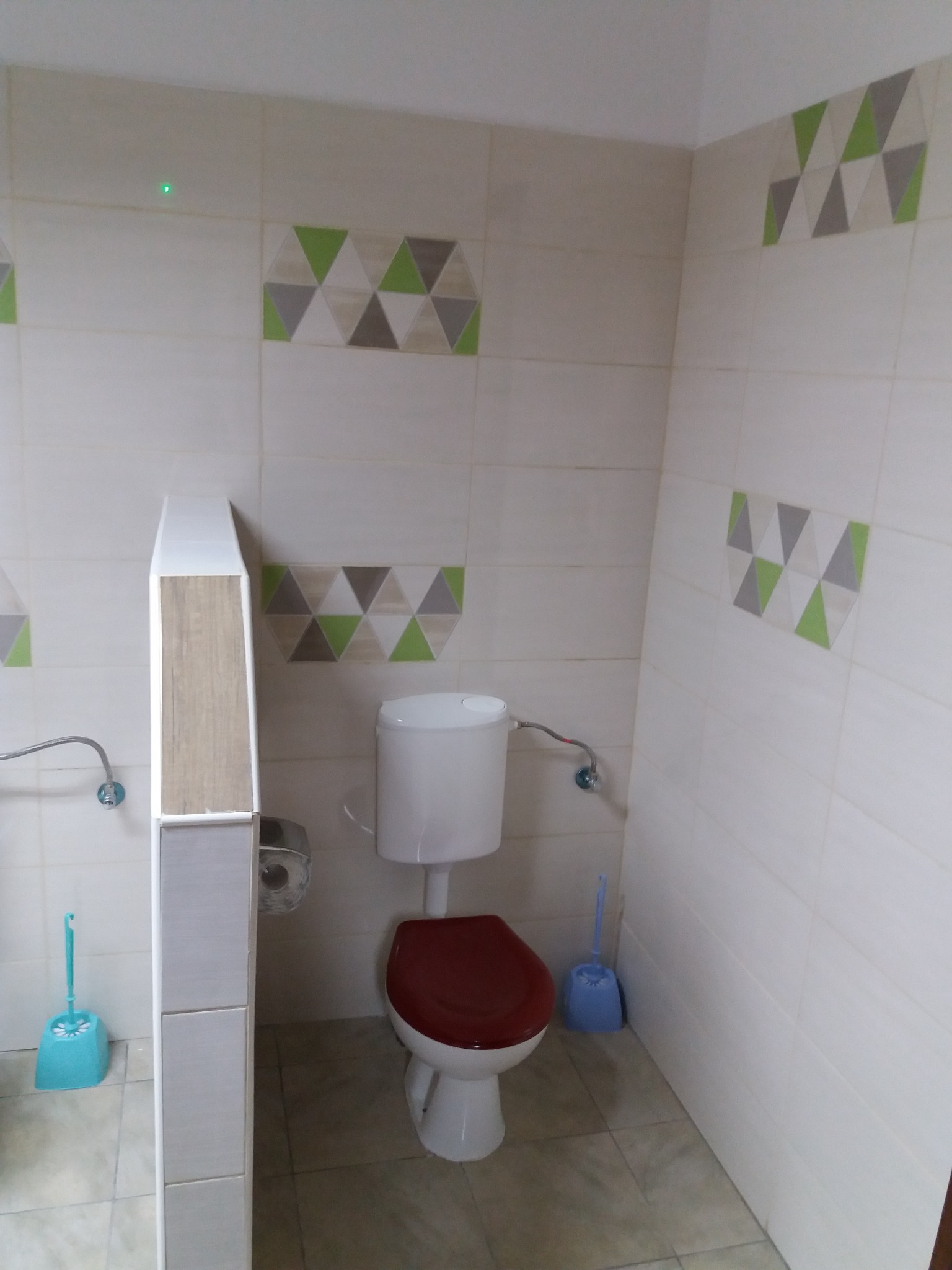 Wykończona łazienka z białymi płytkami, dekoracyjnym pasem z trójkątami w odcieniach szarości i zieleni, toaletą z bordową deską sedesową oraz niebieską szczotką do WC.
