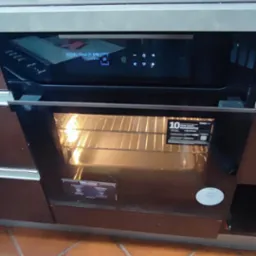 Wbudowany piekarnik z włączonym oświetleniem wewnętrznym, widoczne ruszty, panel sterowania z wyświetlaczem i przyciskami, brązowe płytki podłogowe, fragment ceglanej ściany w tle.