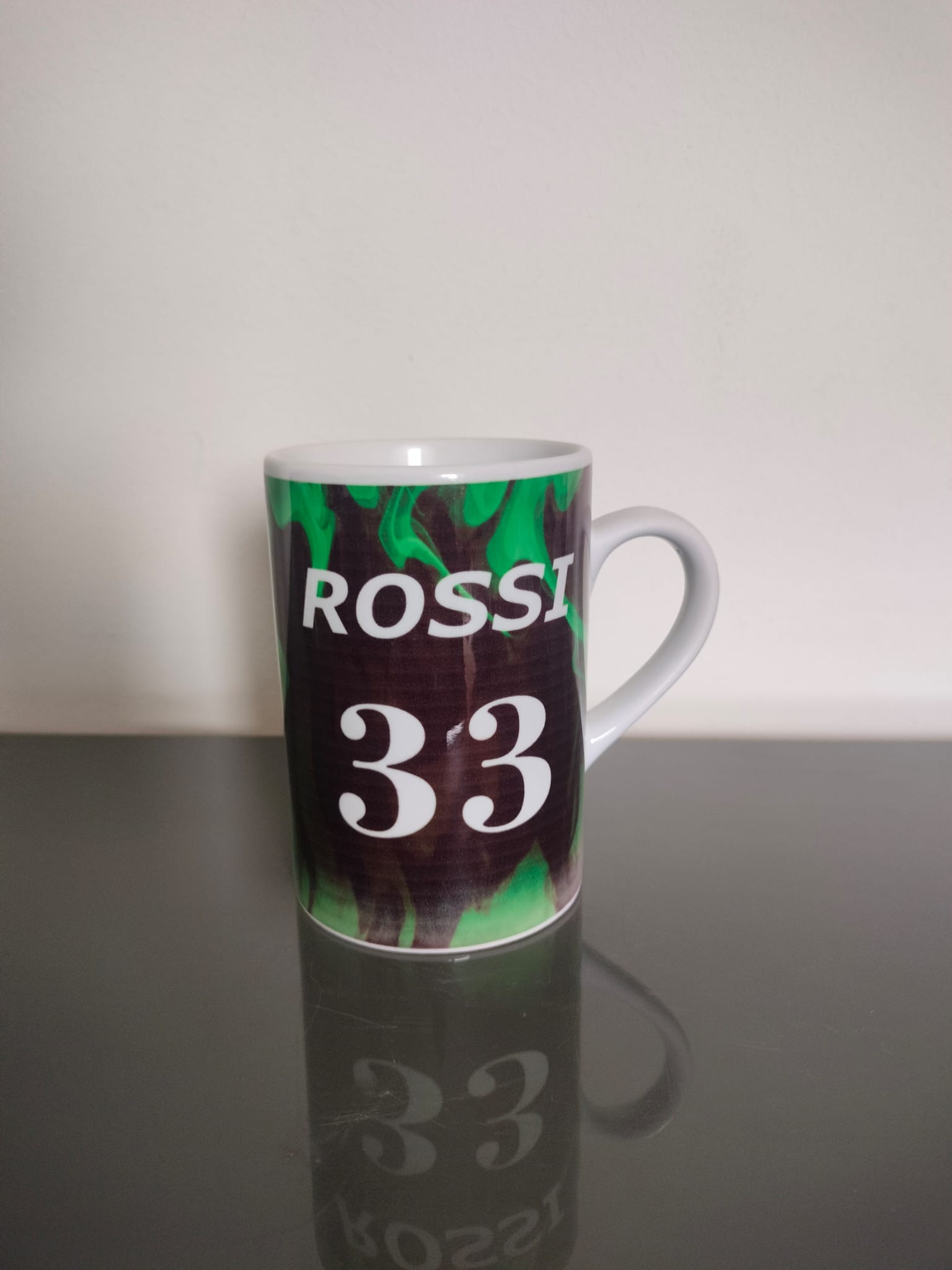 Biały ceramiczny kubek z nadrukiem 'ROSSI 33' na tle zielonych płomieni, stojący na błyszczącej, ciemnej powierzchni, z widocznym odbiciem.