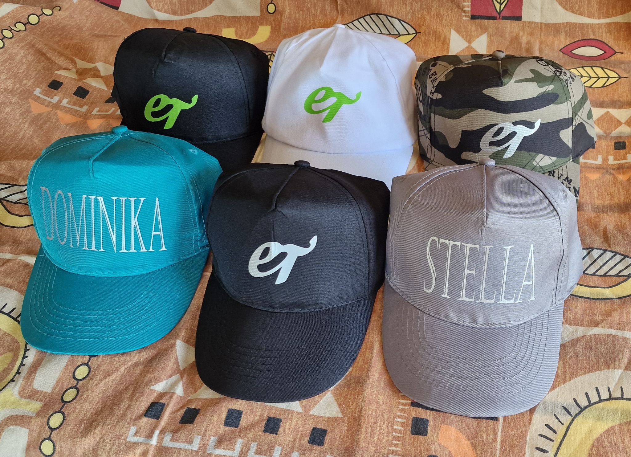 Kolekcja czapek z daszkiem z różnymi nadrukami: imiona (Dominika, Stella) i logo 'et' w różnych kolorach, ułożone na wzorzystej tkaninie.