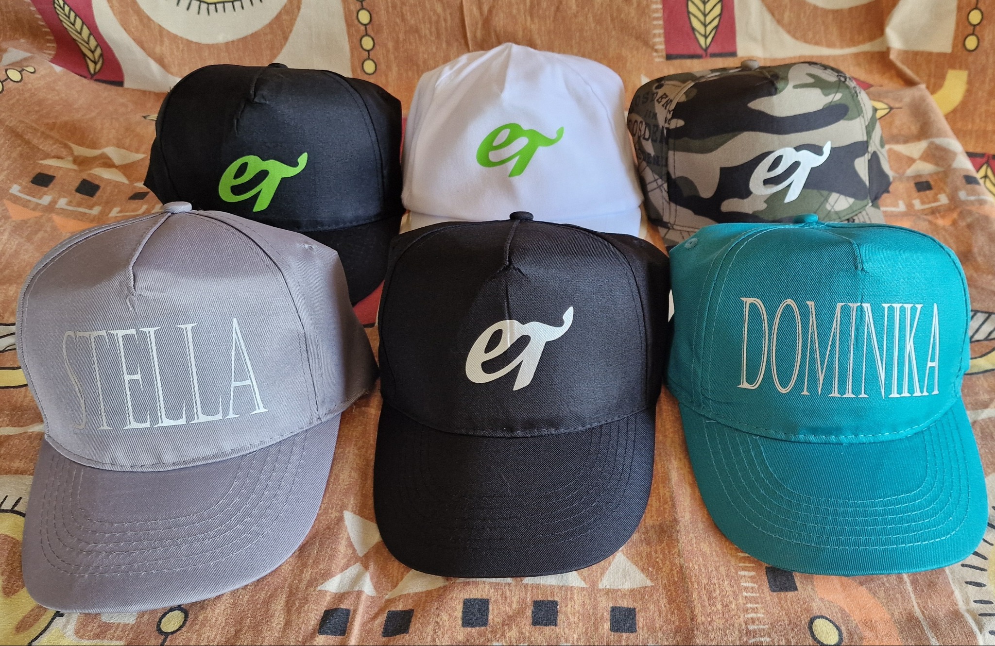 Sześć czapek z daszkiem w różnych kolorach (czarny, biały, szary, zielony, moro) z nadrukami imion i logo, ułożonych na wzorzystym materiale.