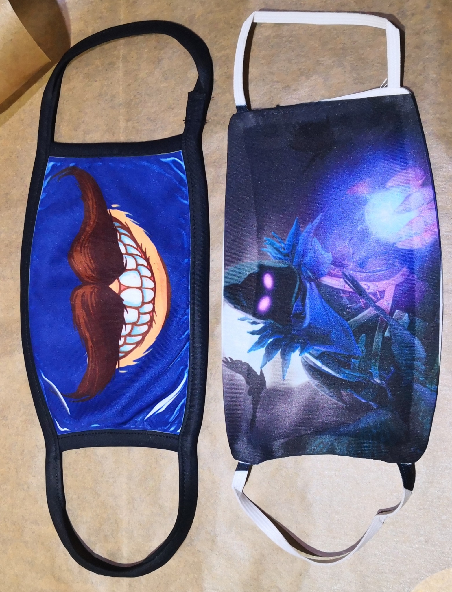 Dwie maseczki ochronne z nadrukami: jedna z motywem uśmiechu z wąsami na niebieskim tle, druga z abstrakcyjnym motywem w odcieniach fioletu i niebieskiego.