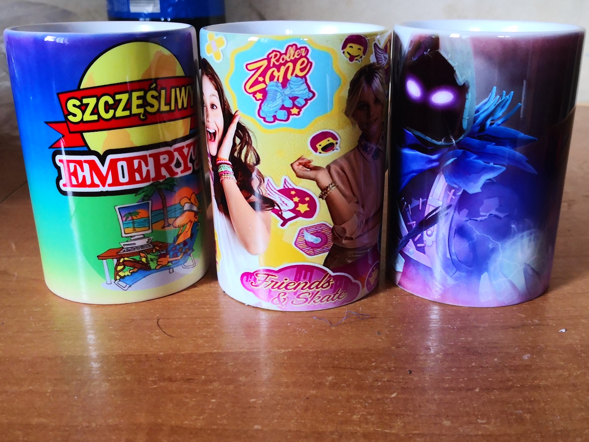 Trzy ceramiczne kubki z różnymi, kolorowymi nadrukami stojące na drewnianym blacie: pierwszy z napisem 'Szczęśliwy Emeryt' i rysunkiem plaży, drugi z motywem 'Roller Zone' i wizerunkami dziewcząt...