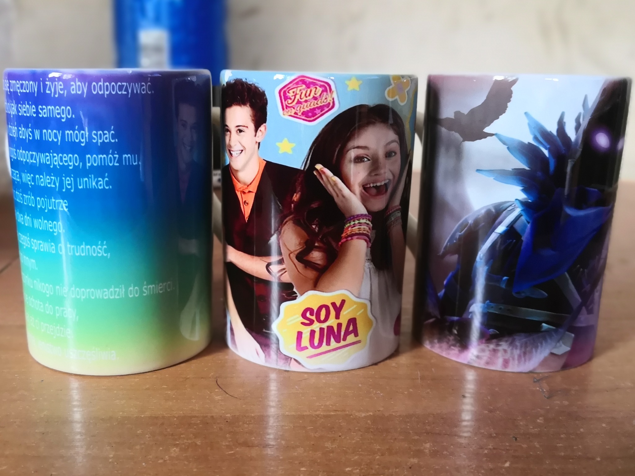 Trzy ceramiczne kubki z różnymi nadrukami: gradientowy z tekstem, z postaciami z serialu 'Soy Luna' i z fantastycznym motywem ptaka i roślin.
