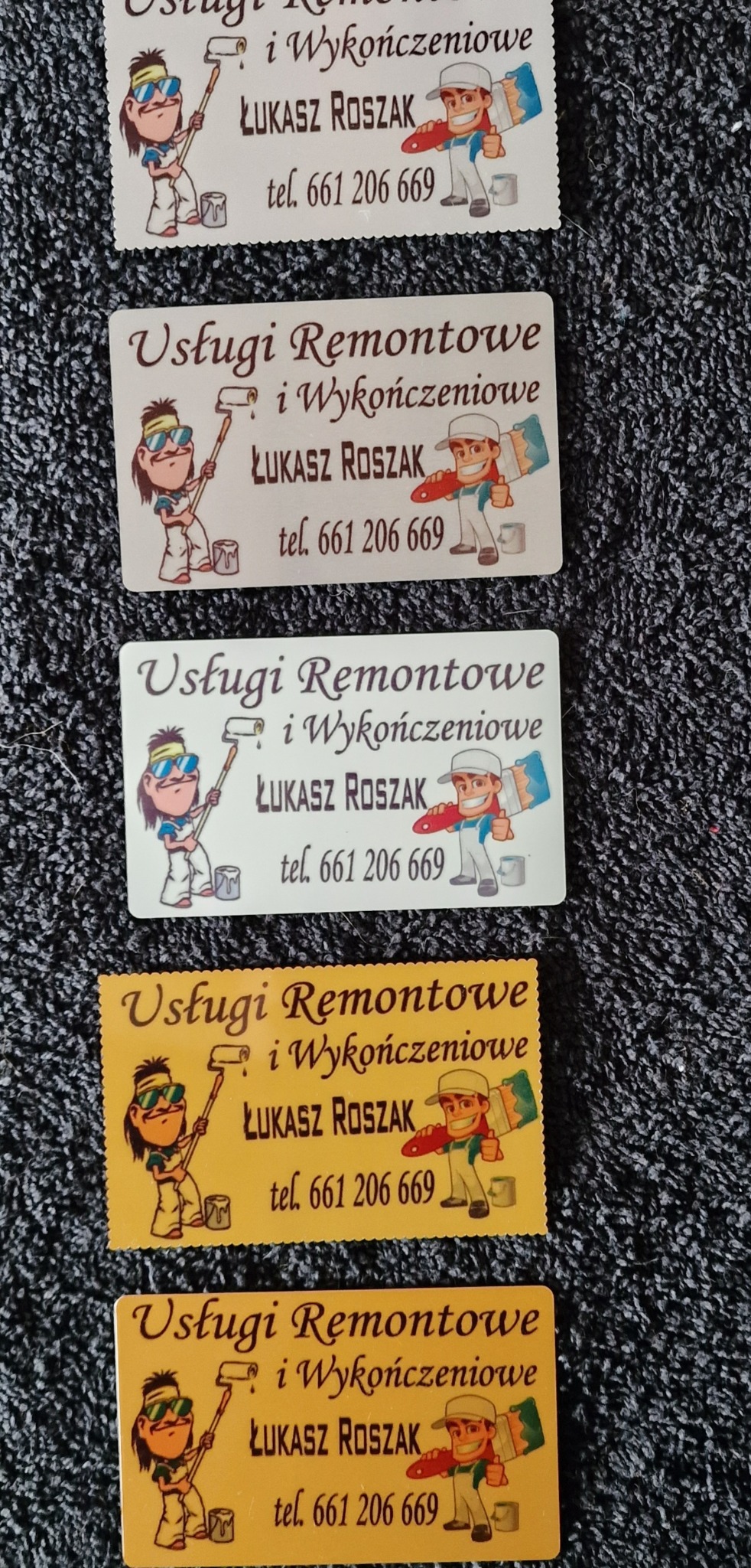Stos wizytówek z rysunkowymi grafikami malarzy i napisem 'Usługi Remontowe i Wykończeniowe Łukasz Roszak tel. 661 206 669' na tle szarej tkaniny.