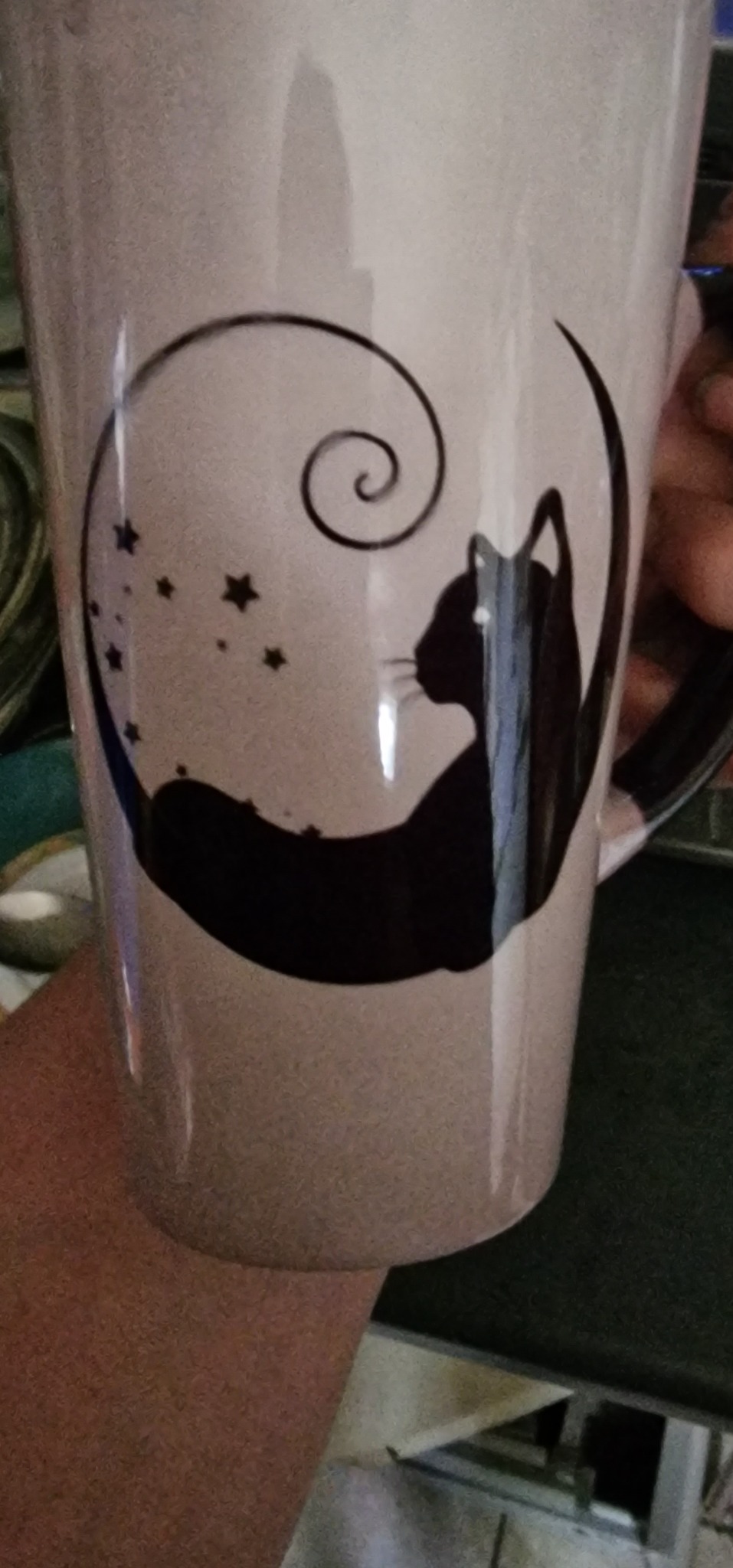 Biały kubek z czarnym nadrukiem przedstawiającym kota siedzącego w półksiężycu z gwiazdami i spiralą.