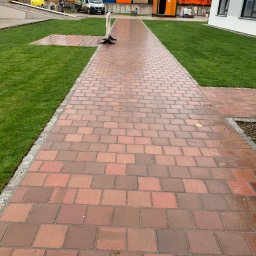 Kostkolada - Czerwona ścieżka z kostki brukowej prowadząca przez trawnik w kierunku budynków, z widocznymi kontenerami budowlanymi i sprzętem budowlanym w tle.