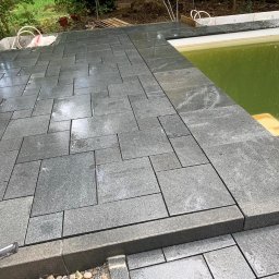 Kostkolada - Świeżo ułożona, mokra, grafitowa nawierzchnia z nieregularnych płyt kamiennych wokół basenu z zielonkawą wodą, widoczne narzędzie do układania kostki.