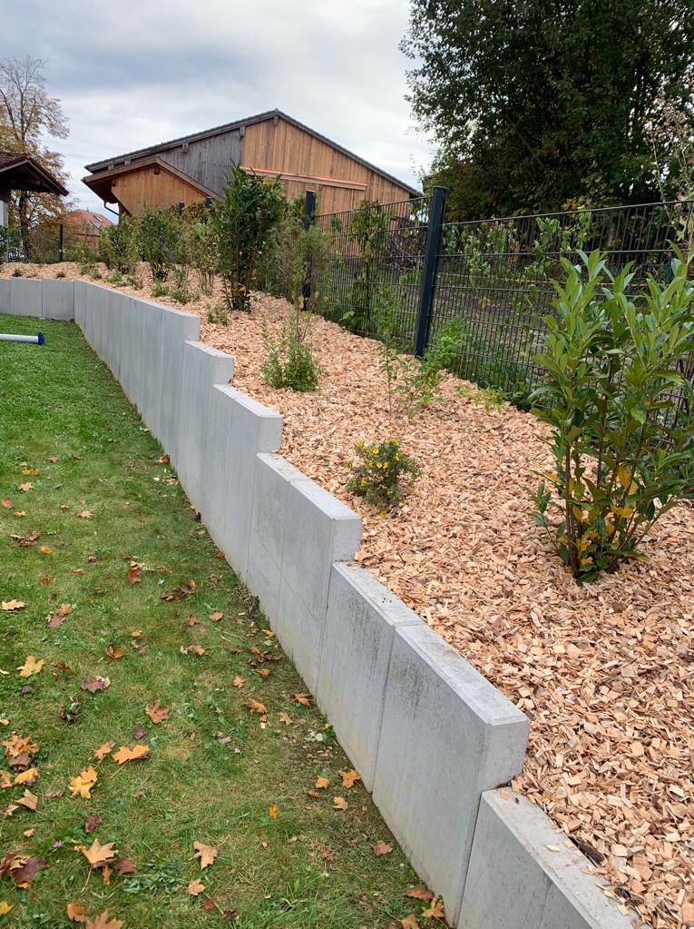 Betonowa ściana oporowa z roślinami i korą drzewną, widok na ogród z domem w tle.