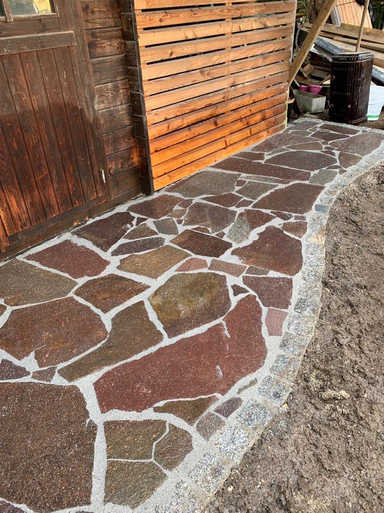 Ścieżka z nieregularnych, brązowo-czerwonych płyt kamiennych, wypełniona jasnym kruszywem, obrzeżona szarym kamieniem, prowadząca obok drewnianej szopy i płotu.