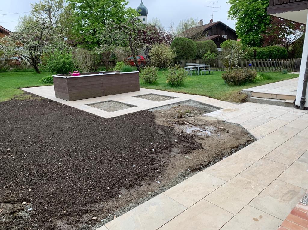 Przygotowanie terenu pod trawnik i rabaty kwiatowe z obrzeżem z jasnej kostki, widoczny fragment tarasu i donica z ciemnego drewna na podwyższeniu.