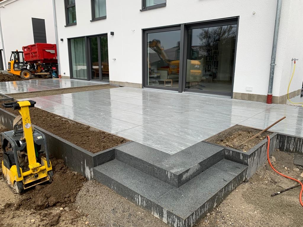 Układanie szarych płytek tarasowych z kamiennymi schodami, ubijarka w tle, widoczny mini-wykopywacz i kontener na gruz, częściowo ukończona realizacja.