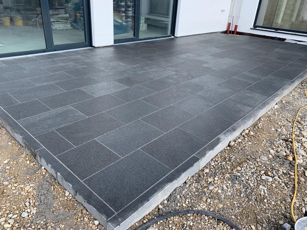 Nowoczesny taras wyłożony ciemnoszarą kostką granitową ułożoną w prostokątnym wzorze, z widocznym obrzeżem i przygotowanym podłożem z kamieni, z przesuwnymi drzwiami balkonowymi w tle.