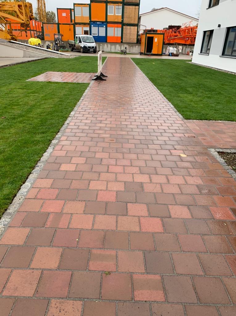 Czerwona ścieżka z kostki brukowej prowadząca przez trawnik w kierunku budynków, z widocznymi kontenerami budowlanymi i sprzętem budowlanym w tle.