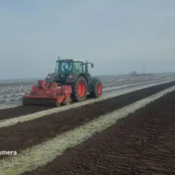 Zielony traktor z czerwonymi kołami i glebogryzarką na polu z rzędami upraw, częściowo pokrytym szronem, w pochmurny dzień.