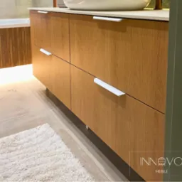 Drewniana szafka łazienkowa z białymi uchwytami, umywalką nablatową i miękkim dywanikiem. Ciepłe oświetlenie podkreśla naturalny odcień drewna.