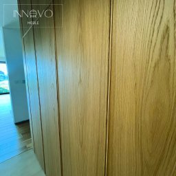 INNOVO MEBLE Magdalena Kłobucka Meble na wymiar - Jasna, drewniana szafa wnękowa z subtelnymi, pionowymi podziałami, dopasowana do wnętrza. Widoczna część korytarza z oknem i zielenią w tle.