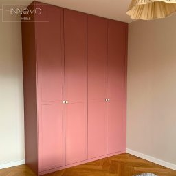 INNOVO MEBLE Magdalena Kłobucka Meble na wymiar - Różowa, czterodrzwiowa szafa z okrągłymi uchwytami na tle parkietu i beżowej ściany, oświetlona lampą. Minimalistyczny design, gładkie fronty.