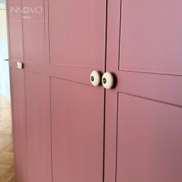 INNOVO MEBLE Magdalena Kłobucka Meble na wymiar - Różowa szafa z kremowymi gałkami w stylu retro. Detal wykończenia, widoczne łączenia frontów i delikatna faktura farby. Parkiet na podłodze.