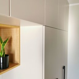 INNOVO MEBLE Magdalena Kłobucka Meble na wymiar - Jasna szafa wnękowa z drewnianą półką i rośliną w czarnej doniczce. Minimalistyczny design z długimi, czarnymi uchwytami.