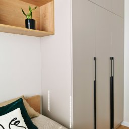 INNOVO MEBLE Magdalena Kłobucka Meble na wymiar - Beżowa szafa wnękowa z długimi, czarnymi uchwytami w nowoczesnej sypialni. Nad szafą drewniana półka z zieloną rośliną. Częściowo widoczne łóżko.