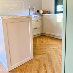 INNOVO MEBLE Magdalena Kłobucka Meble na wymiar - Jasna, narożna kuchnia z frezowanymi frontami i marmurowym blatem, widok na parkiet ułożony w jodełkę, czarne uchwyty, minimalistyczny plakat na blacie.