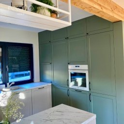 INNOVO MEBLE Magdalena Kłobucka Meble na wymiar - Nowoczesna kuchnia w odcieniach zieleni z białym piekarnikiem i marmurowym blatem wyspy, doświetlona naturalnym światłem z okna z roletą.