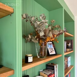 INNOVO MEBLE Magdalena Kłobucka Meble na wymiar - Zielona, drewniana biblioteczka z dębowymi półkami, na których stoją książki, świeca i wazon z suchymi kwiatami. Ujęcie z bliska, widać detale wykończenia.